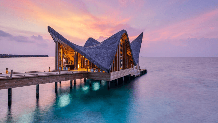 Overwater pavilion with a wave-shaped roof at sunset