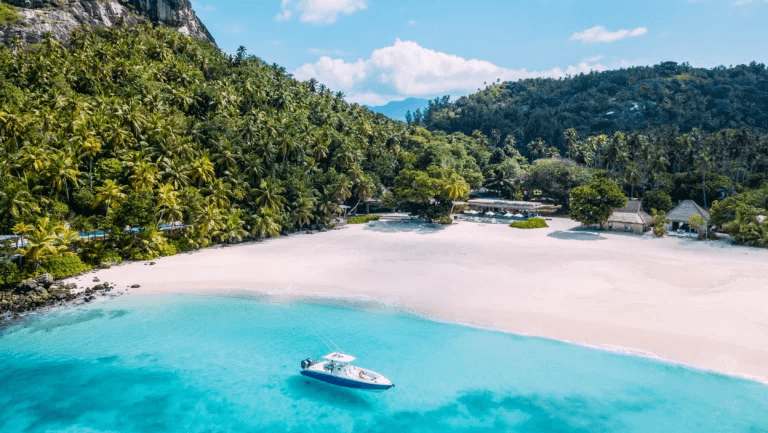 Boat in turquoise water off a wide white beach below forested hills