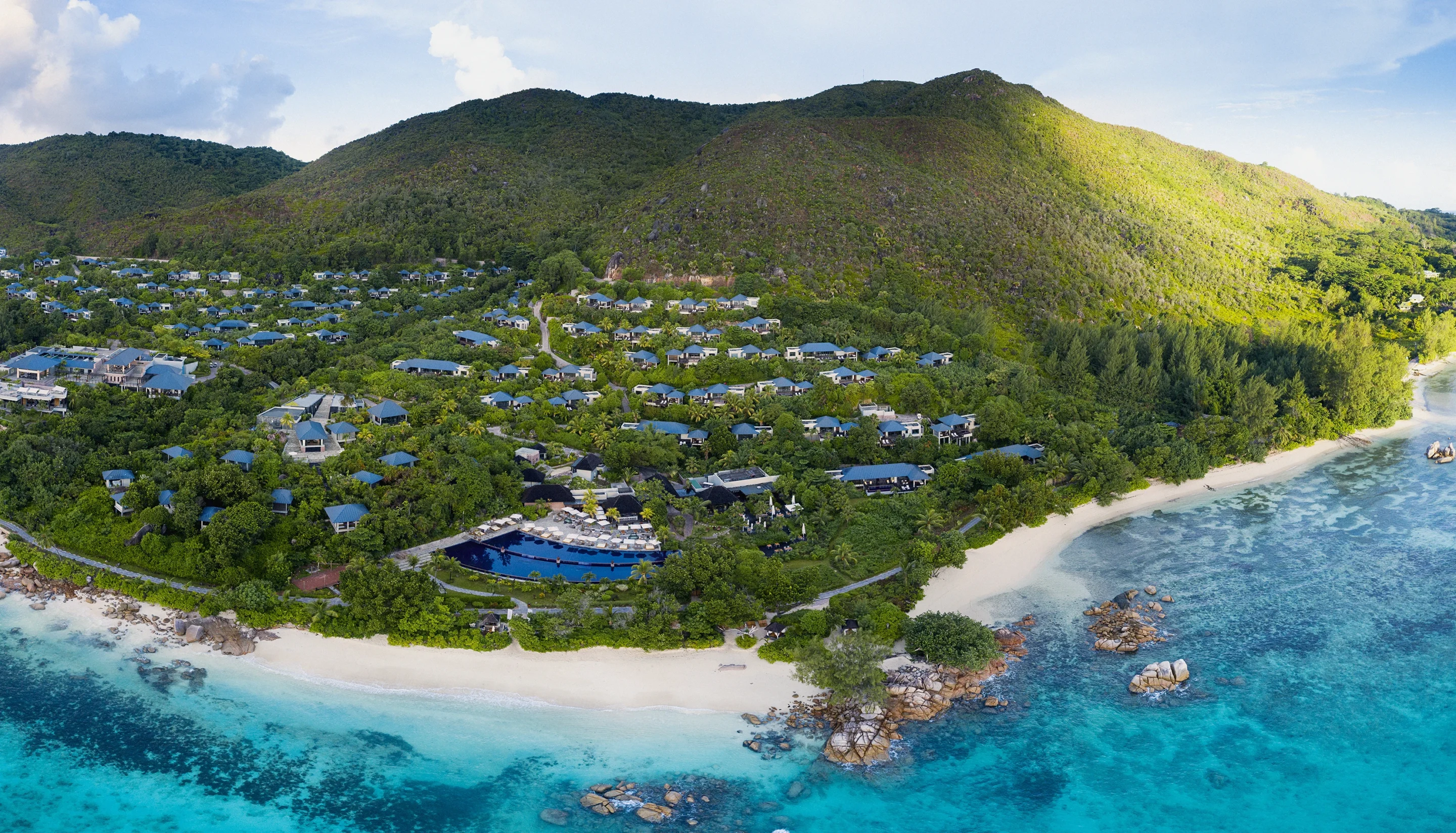 Villas spread across a green hillside above a white beach, seen from the air