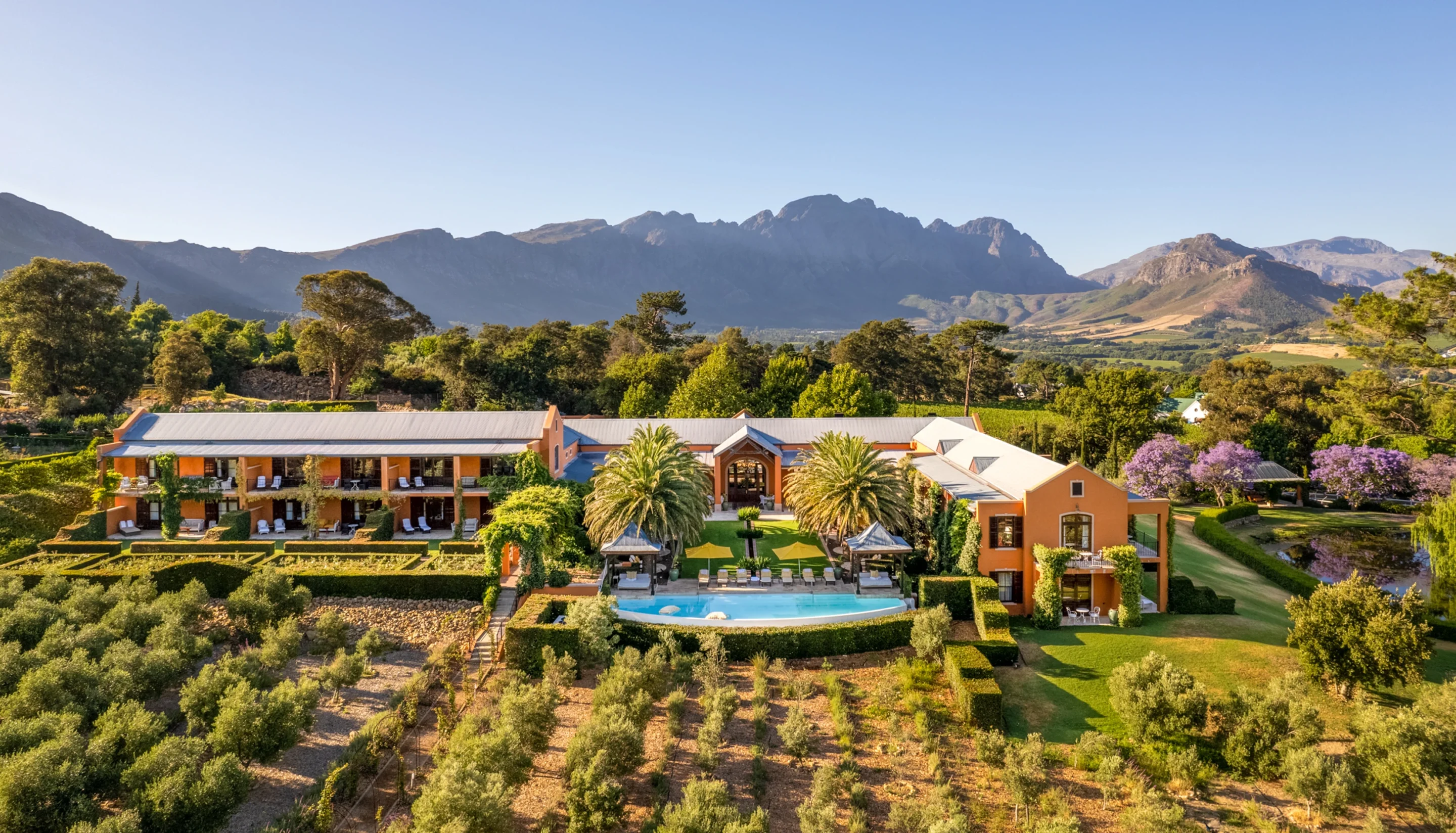 Hotel and pool among orchards below mountains, seen from the air