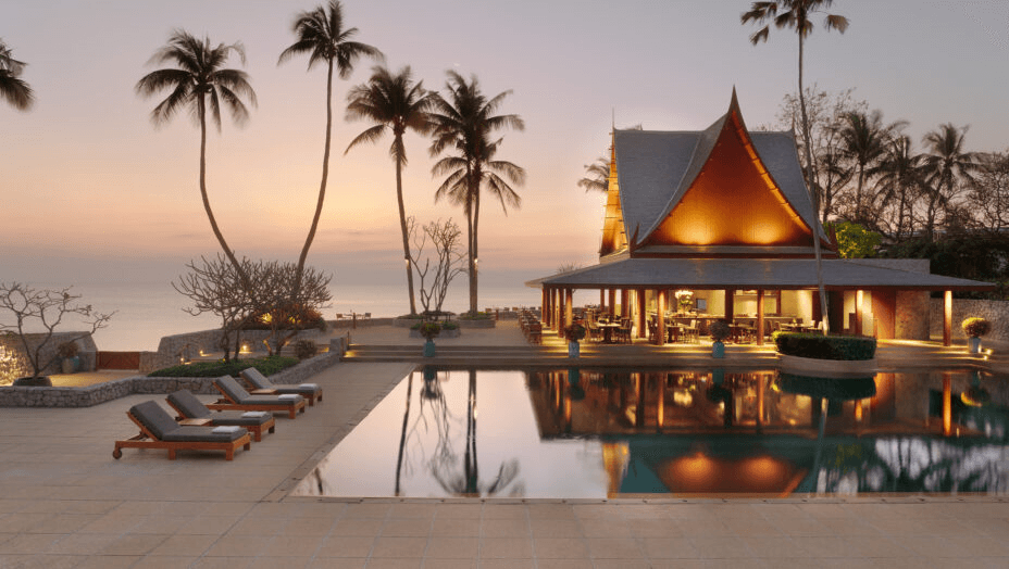 Thai pavilion lit at dusk beside a pool and palms