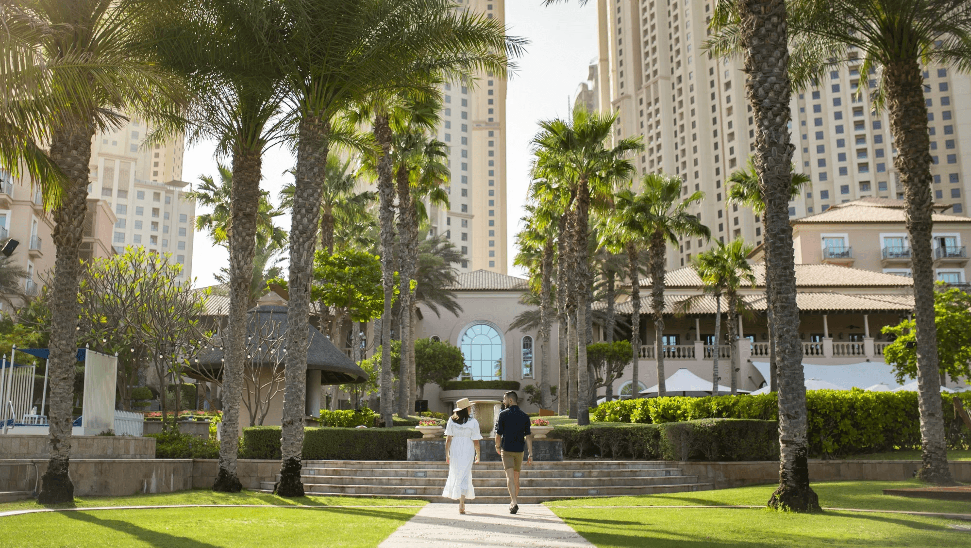 Couple walking along a palm-lined garden path towards the hotel, with towers behind