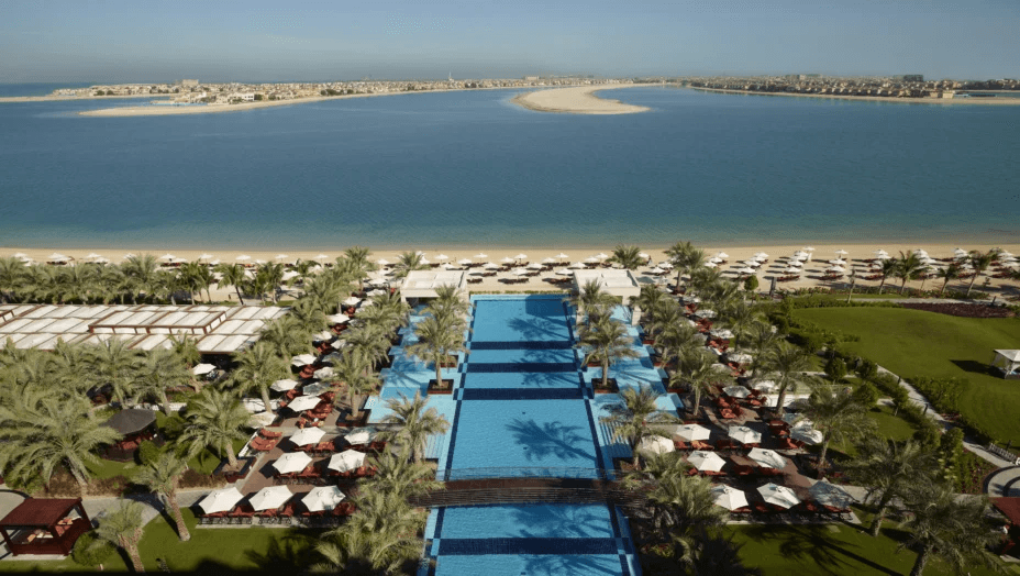 Long pool between palms and sunshades, with the sea beyond, seen from above